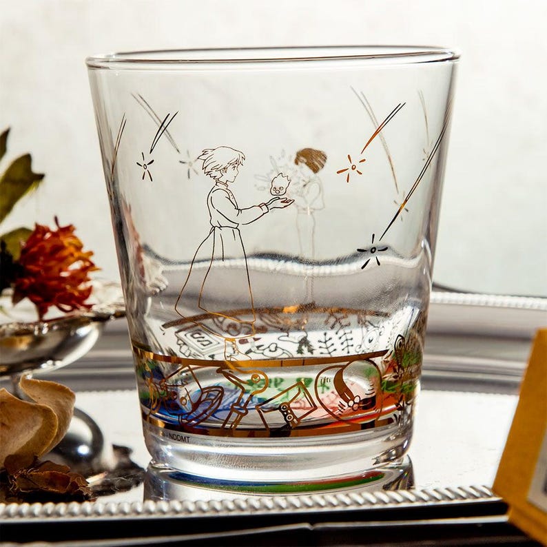 May include: A clear glass tumbler with a gold-colored illustration of a character from an animated film. The design features a character holding a small creature, with shooting stars and a decorative band at the base. The glass is on a silver tray.