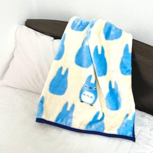 May include: A cream-colored plush blanket featuring a pattern of blue cartoon characters. The blanket has a dark blue border and is draped over a white pillow and bed. The blanket is soft and cozy.