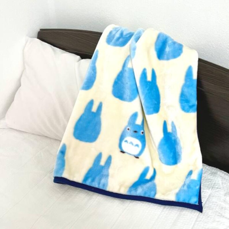 May include: A cream-colored plush blanket featuring a pattern of blue cartoon characters. The blanket has a dark blue border and is draped over a white pillow and bed. The blanket is soft and cozy.