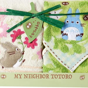 以下が含まれることがあります： 2枚の「となりのトトロ」をテーマにしたタオルセット。1枚は花を持つ茶色のウサギ、もう1枚は青いトトロのキャラクターが特徴です。セットは緑のリボンで結ばれ、タグが付いています。