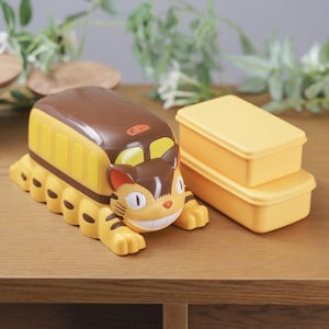 Może przedstawiać: Żółto-brązowy pojemnik na lunch w stylu kreskówki w kształcie autobusu-kota, z uśmiechniętą twarzą i paskami. Obok znajdują się dwa żółte prostokątne pojemniki na lunch, wszystko na drewnianej powierzchni.