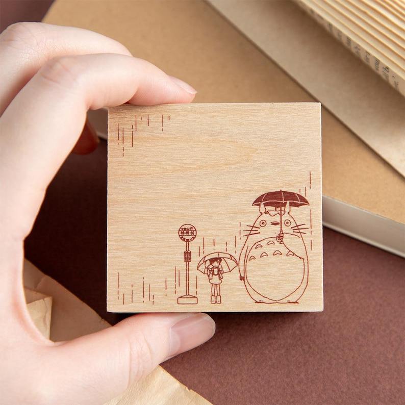 Op de afbeelding: Een vierkante houten rubberstempel met een bruine lijntekening van een groot wezen en een klein figuurtje die onder paraplu's in de regen bij een bushalte staan. Het ontwerp bevat verticale lijnen die de regen voorstellen.