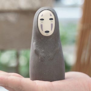 Puede incluir: Una peque&ntilde;a figura de color gris oscuro con un cuerpo liso y redondeado y una cara p&aacute;lida de forma ovalada. La cara presenta dos ojos negros horizontales, una boca negra peque&ntilde;a y dos marcas verticales moradas bajo los ojos.