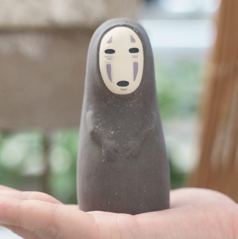 Puede incluir: Una peque&ntilde;a figura de color gris oscuro con un cuerpo liso y redondeado y una cara p&aacute;lida de forma ovalada. La cara presenta dos ojos negros horizontales, una boca negra peque&ntilde;a y dos marcas verticales moradas bajo los ojos.