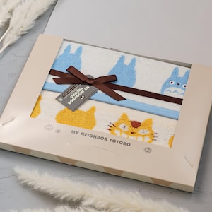 May include: A gift set featuring two white towels with blue and yellow cartoon character designs, tied with a brown ribbon. The box has the text "MY NEIGHBOR TOTORO".