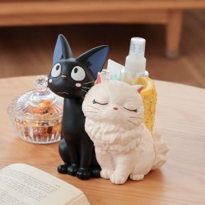 Puede incluir: Un organizador de escritorio con una figura de gato negro de orejas grandes y una figura de gato blanco esponjoso con los ojos cerrados. Detr&aacute;s hay un recipiente amarillo texturizado con un atomizador y un tubo de crema blanco.