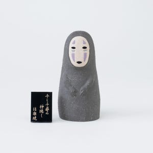 Puede incluir: Una figura de cer&aacute;mica de color gris oscuro texturizado que representa un personaje con rostro blanco p&aacute;lido, marcas moradas y ojos negros. Junto a la figura hay una peque&ntilde;a placa rectangular negra con texto japon&eacute;s dorado: 千と千尋の神隠し 銘楽焼.