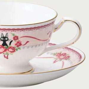 May include: White teacup and saucer set with a gold trim. The teacup features a black cat illustration, red roses, and a pink lace design. The saucer has a matching floral pattern.