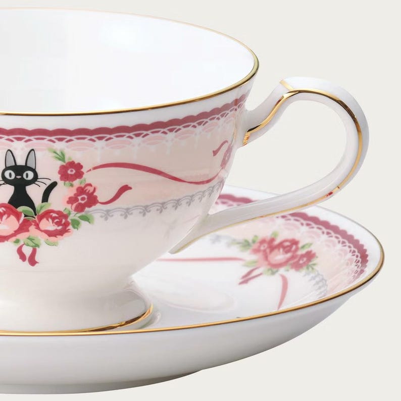 May include: White teacup and saucer set with a gold trim. The teacup features a black cat illustration, red roses, and a pink lace design. The saucer has a matching floral pattern.