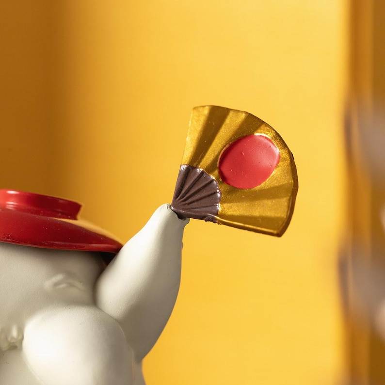Peut inclure: Une figurine blanche avec un chapeau rouge et un petit &eacute;ventail dor&eacute; avec un cercle rouge au centre. L'&eacute;ventail est lev&eacute; en l'air. Le fond est de couleur jaune uni.