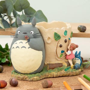 Puede incluir: Una maceta caprichosa con personajes de la película de animación "Mi vecino Totoro". La maceta tiene un diseño de tronco de árbol beige claro con una figura de Totoro gris sosteniendo una bellota. También aparece una niña con un vestido rosa y un Totoro azul.