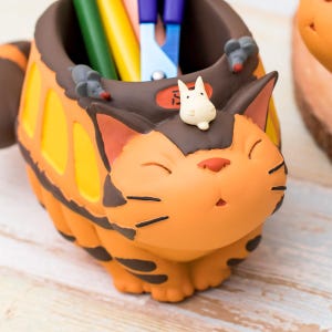May include: An orange and brown cat-shaped desk organizer with a smiling face, black whiskers, and a white bunny figure on top. The organizer has a bus-like design with yellow windows and holds pens and scissors. Two gray mouse figures are on the rim.