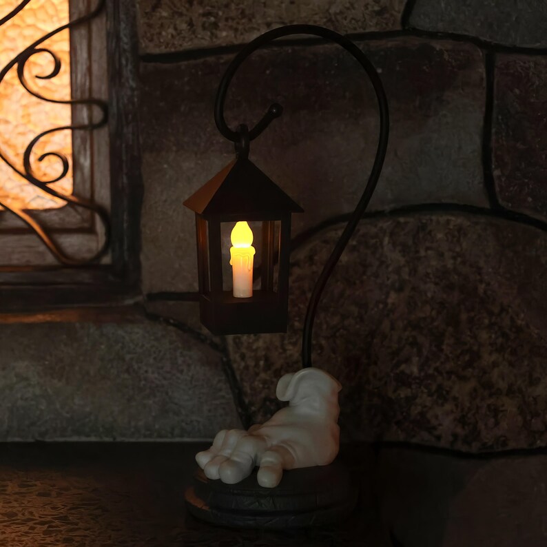 May include: A decorative black metal lantern with a curved handle, featuring a flickering candle-shaped light. The base is a white sculpture of a hand resting on a dark, round platform. The lantern is set against a stone wall.