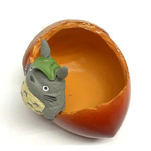 K&ouml;nnte beinhalten: Ein dekorativer eichelf&ouml;rmiger Beh&auml;lter mit einer herausblickenden Zeichentrickfigur. Der Beh&auml;lter ist haupts&auml;chlich orange und braun, mit glattem Innenraum. Die Figur ist grau mit einem gr&uuml;nen Hut und einem gelb gemusterten Bauch.
