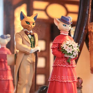 Puede incluir: Figuras de gatos antropom&oacute;rficos vestidos con atuendos formales. Un gato lleva un traje beige con chaleco verde y bast&oacute;n. El otro gato viste un vestido rojo y un sombrero, sosteniendo un ramo de flores blancas. Las figuras tienen caras de gato detalladas.