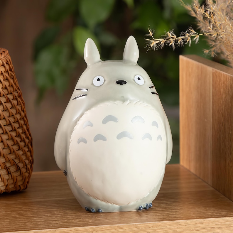 Puede incluir: Una figura gris y blanca de Totoro, un personaje de la pel&iacute;cula de animaci&oacute;n Mi Vecino Totoro. La figura tiene ojos grandes, orejas puntiagudas y una barriga blanca con marcas en forma de media luna. Se exhibe en una estanter&iacute;a de madera.