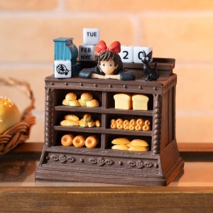 Pode incluir: Uma cena de padaria em miniatura detalhada com uma jovem com um la&ccedil;o vermelho, um gato preto e v&aacute;rios produtos de panifica&ccedil;&atilde;o. A cena inclui um calend&aacute;rio mostrando "TUE FEB 2". A padaria &eacute; marrom com prateleiras de p&atilde;o e pastelaria.