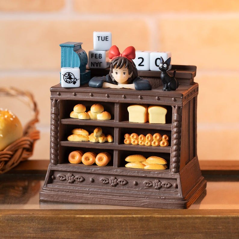 Pode incluir: Uma cena de padaria em miniatura detalhada com uma jovem com um la&ccedil;o vermelho, um gato preto e v&aacute;rios produtos de panifica&ccedil;&atilde;o. A cena inclui um calend&aacute;rio mostrando "TUE FEB 2". A padaria &eacute; marrom com prateleiras de p&atilde;o e pastelaria.