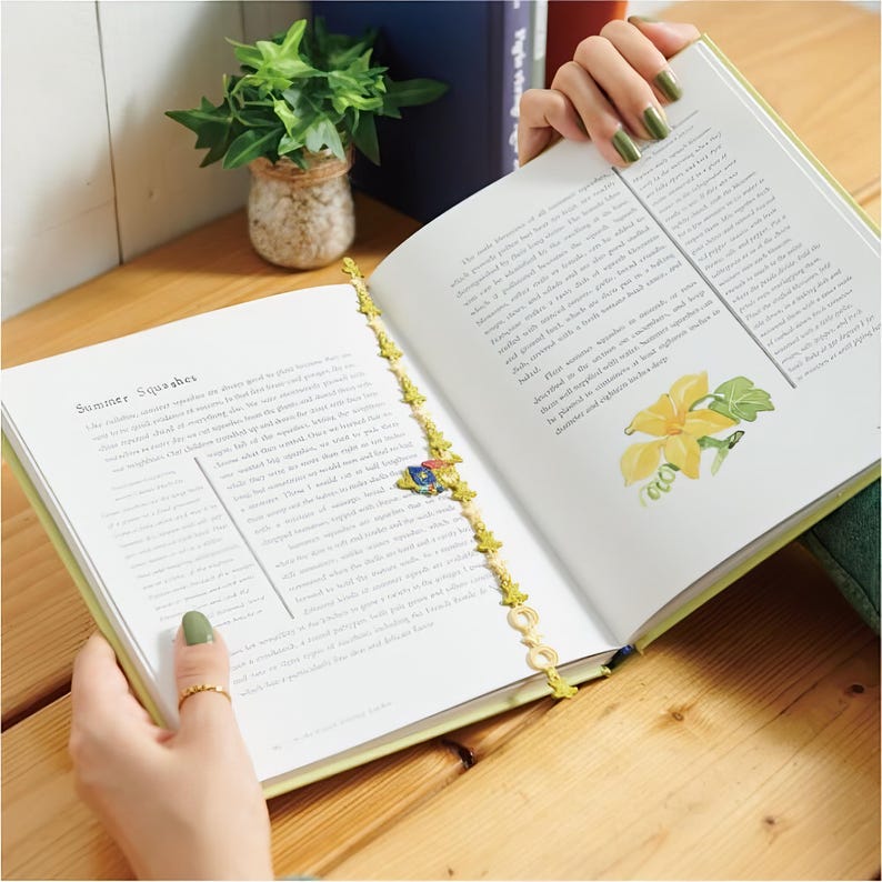 K&ouml;nnte beinhalten: Ein offenes Buch mit dem Text "Summer Squashes" und einem dekorativen goldfarbenen Lesezeichen. Das Lesezeichen hat kleine, blattf&ouml;rmige Anh&auml;nger und einen zentralen Anh&auml;nger mit blauem, rotem und gelbem Design. Das Buch liegt auf einer Holzoberfl&auml;che.