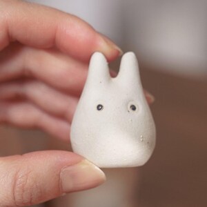 May include: A small, white ceramic figurine with a rounded body and two upright ears. The figurine has two small, dark eyes and a smooth, matte finish. The object is held in a person's hand.