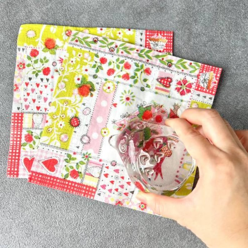 May include: Two decorative floral napkins with a clear glass teapot. The napkins feature a patchwork design with strawberries, hearts, and floral patterns in red, pink, and green. The teapot is held by a hand.