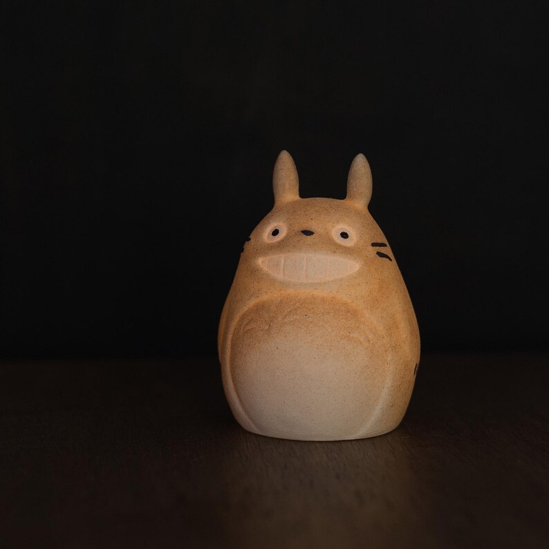 Może przedstawiać: Jasnobeżowa, zaokrąglona ceramiczna figurka w kształcie stworzenia z dwoma spiczastymi uszami, dużymi oczami, szerokim uśmiechniętym pyskiem i trzema małymi wąsami po każdej stronie twarzy. Powierzchnia ma matowe wykończenie z delikatnym cieniowaniem.