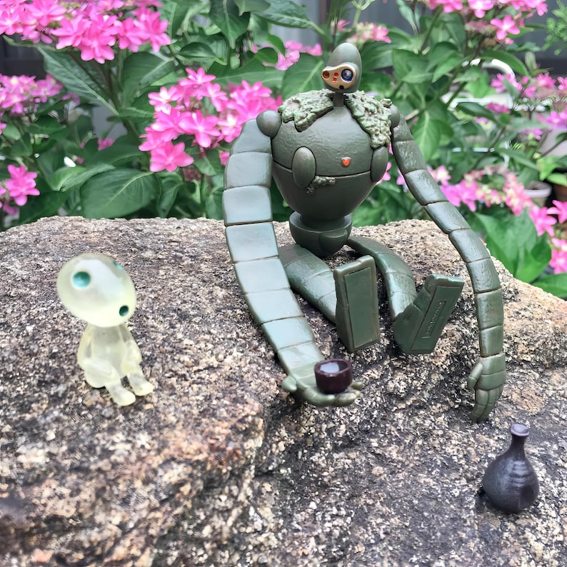 Peut inclure: Une figurine de robot vert avec des membres articul&eacute;s est assise sur un rocher, tenant une petite tasse. Une figurine translucide et brillante est assise &agrave; proximit&eacute;. Une petite bouteille sombre repose sur le rocher. Des fleurs roses fleurissent en arri&egrave;re-plan.