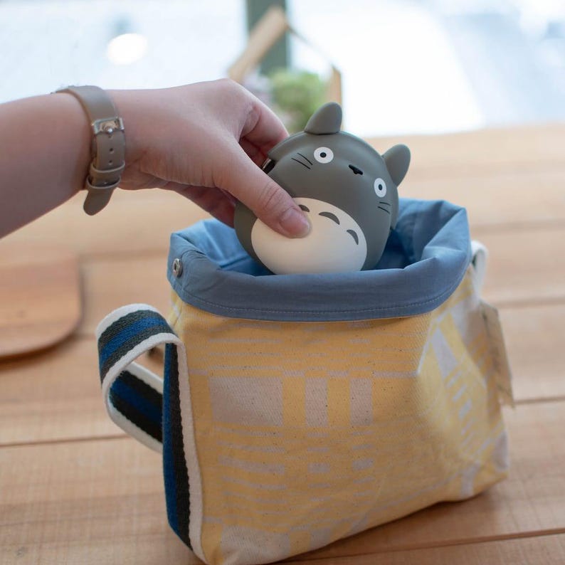 May include: A gray Totoro figure with white belly detail is placed inside a yellow canvas bag with a blue interior. The bag has a striped handle with blue and black accents. The bag is on a wooden surface.