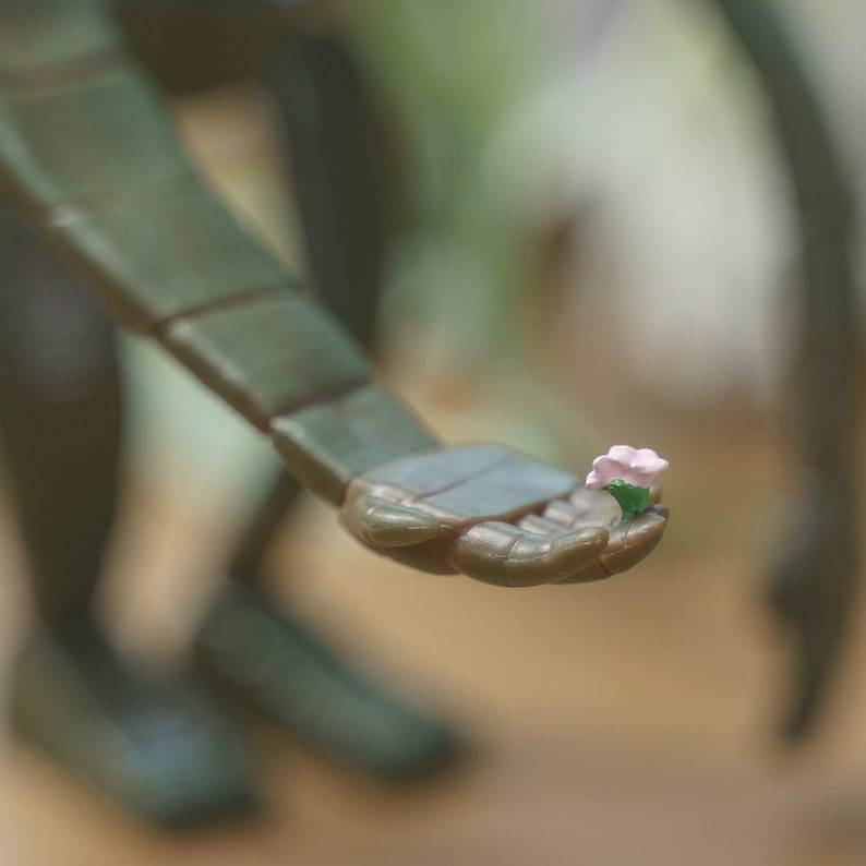 Peut inclure: Gros plan d'un bras vert articul&eacute; tenant une petite fleur rose avec des feuilles vertes. Le bras est segment&eacute; et semble faire partie d'une figure plus grande. L'arri&egrave;re-plan est flou, sugg&eacute;rant un cadre ext&eacute;rieur.