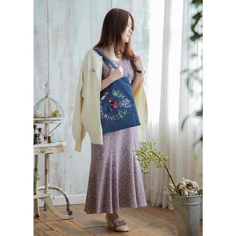 K&ouml;nnte beinhalten: Eine Frau tr&auml;gt einen wei&szlig;en Cardigan und ein gemustertes Kleid und h&auml;lt eine blaue Tragetasche mit einem gestickten Blumendesign. Die Tasche hat einen roten Vogel und gr&uuml;ne Bl&auml;tter.