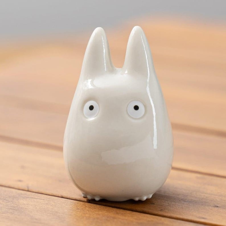 K&ouml;nnte beinhalten: Kleine, cremefarbene Keramikfigur einer Zeichentrickfigur mit gro&szlig;en, runden Augen und spitzen Ohren. Die Figur hat eine glatte, gl&auml;nzende Oberfl&auml;che und steht auf einer Holzoberfl&auml;che. Das Design der Figur ist einfach und minimalistisch.