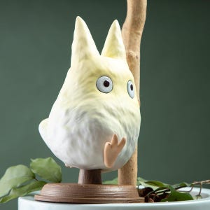 May include: A whimsical, cream-colored figurine of a cartoon character with large, expressive eyes and pointed ears. The figure has a rounded body and is perched on a brown, circular base. A small, tan hand is visible. The background is a muted green.