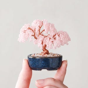 Op de afbeelding: Een miniatuur bonsai boom met een roze kralenluifel en een koperkleurige stam. De boom staat in een kleine, donkerblauwe keramische pot gevuld met kleine witte steentjes. De bonsai wordt tussen twee vingers gehouden.