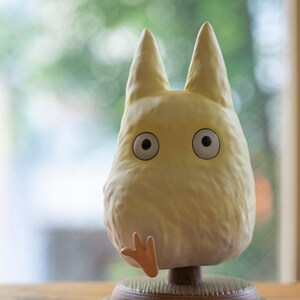 May include: A light yellow figurine of a cartoon character with large, round eyes and pointed ears. The figurine has a small, outstretched hand and is mounted on a brown base. The background is blurred, with a window and greenery visible.