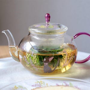 Puede incluir: Una tetera de cristal transparente con asa y pomo de tapa de color morado. El cuerpo est&aacute; decorado con un gr&aacute;fico de un peque&ntilde;o gato negro con orejas grandes y un lazo rosa, rodeado de ramitas de flores amarillas y moradas.