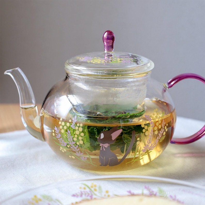 Puede incluir: Una tetera de cristal transparente con asa y pomo de tapa de color morado. El cuerpo est&aacute; decorado con un gr&aacute;fico de un peque&ntilde;o gato negro con orejas grandes y un lazo rosa, rodeado de ramitas de flores amarillas y moradas.