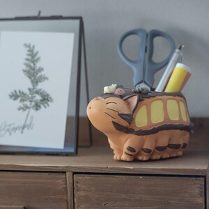 May include: A tan and brown cartoon-style desk organizer shaped like a bus with a cat-like face. The organizer holds gray scissors, a pen, and a yellow cylindrical object. A framed botanical print is in the background.