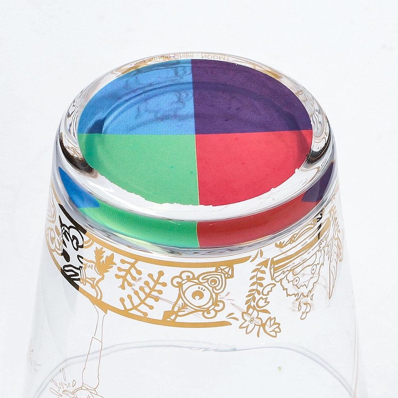 May include: Clear glass with a colorful, segmented base featuring blue, purple, green, and red squares. The glass has a gold decorative border with a bear and floral design. The glass is likely for drinking.