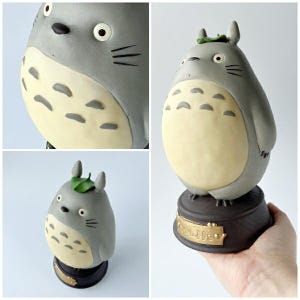 May include: A gray and cream-colored Totoro figurine with a leaf on its head. The figurine has a round belly and is mounted on a dark brown base with gold lettering. The figurine is a collectible item.