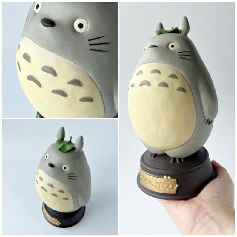 May include: A gray and cream-colored Totoro figurine with a leaf on its head. The figurine has a round belly and is mounted on a dark brown base with gold lettering. The figurine is a collectible item.