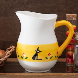 May include: A white ceramic pitcher featuring a wide horizontal yellow band around the center. The band is decorated with a repeating pattern of small red flowers and a black cat illustration. The pitcher has a curved yellow handle and a wide spout.