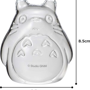 May include: A clear glass figurine in the shape of a cartoon character, 8.5 cm tall and 8.5 cm wide. The character has large eyes, a round belly, and pointed ears. The Studio Ghibli logo is printed on the bottom.