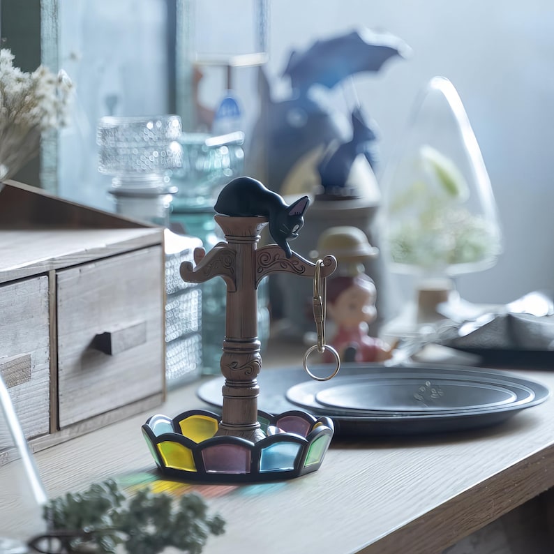 May include: A decorative key holder featuring a black cat figurine perched atop a brown, ornate stand. The base of the stand is a colorful stained-glass design. A set of keys hangs from the holder. The holder sits on a wooden surface.