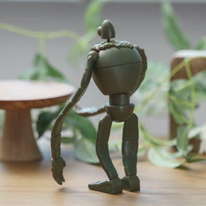 Peut inclure: Une figurine de robot articul&eacute; vert fonc&eacute; avec un corps arrondi, de longs bras et des jambes segment&eacute;es. La figurine a une petite t&ecirc;te et un col en mati&egrave;re textur&eacute;e. Le robot est pos&eacute; sur une surface en bois.