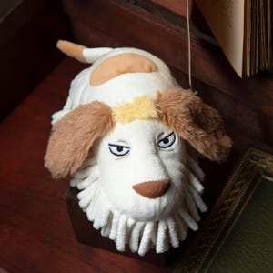 May include: A plush dog toy with a white body, brown ears, and a tan spot. The dog has embroidered eyes and a brown nose. The toy is lying on a dark brown surface.