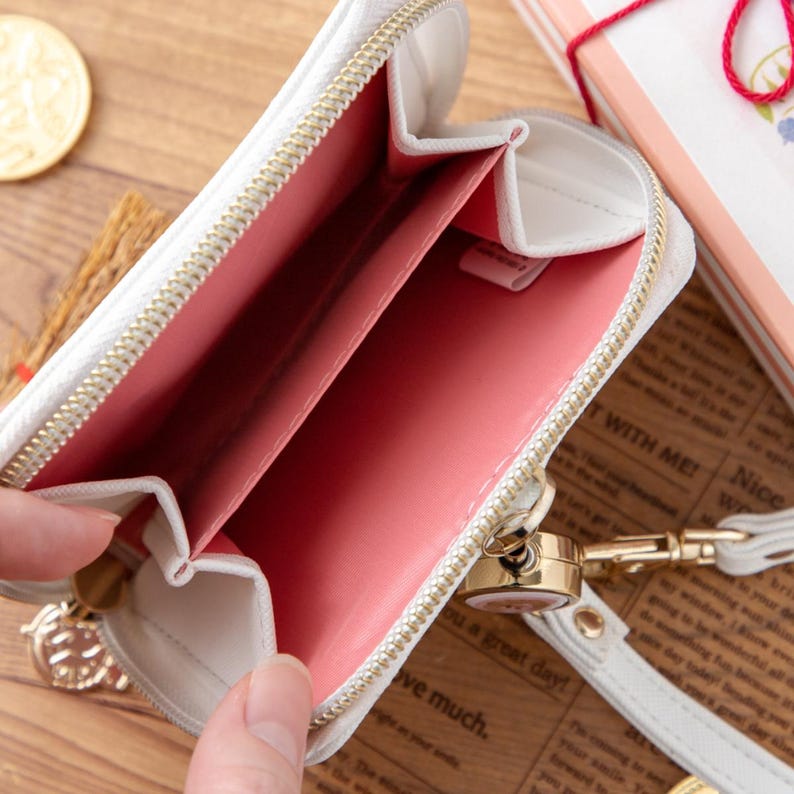 May include: White zippered wallet with a pink interior. The wallet is open, revealing the interior compartments. A gold-colored zipper and a wrist strap are visible. The wallet is on a wooden surface.