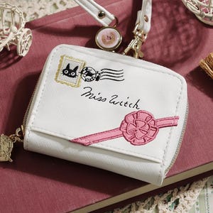 May include: White wallet with a zipper closure, featuring an embroidered design of a black cat, a postage stamp, and the text "Miss Witch." A pink ribbon is also embroidered on the wallet. A small broom and a decorative bicycle are also visible.