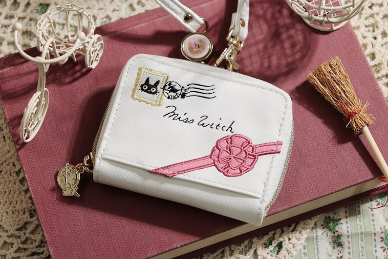 May include: White wallet with a zipper closure, featuring an embroidered design of a black cat, a postage stamp, and the text "Miss Witch." A pink ribbon is also embroidered on the wallet. A small broom and a decorative bicycle are also visible.