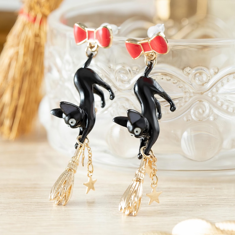 May include: A pair of black cat earrings with red bow accents. The cats hang from gold-colored brooms and star charms. The earrings are set against a blurred background, showcasing a whimsical and playful design.