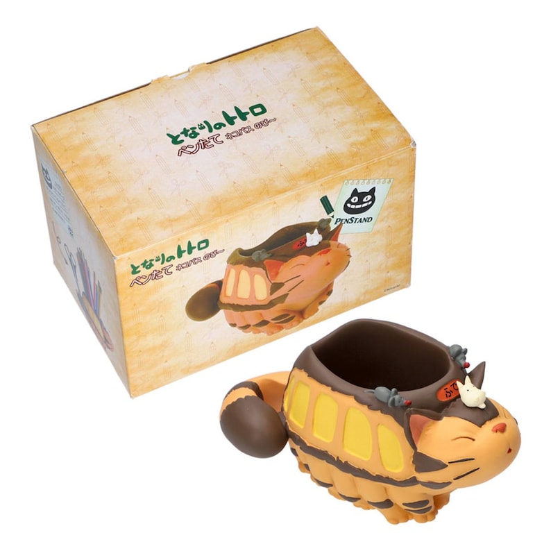 May include: A tan and yellow pen stand shaped like a cat-like creature with a brown tail and accents. The pen stand is next to a box with Japanese text and a cartoon image of the pen stand. The box has a sticker that says "PINSTAND".